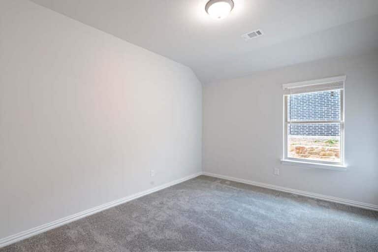 An empty room with gray carpeting and a single window with a view of a brick exterior.