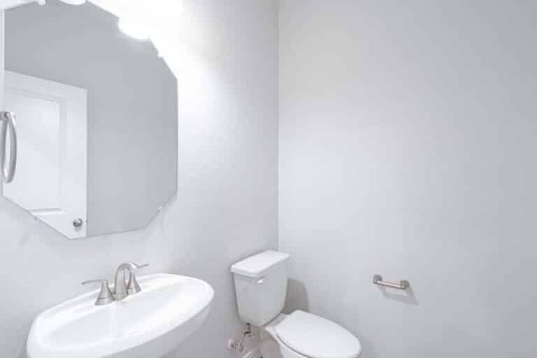 A neatly kept bathroom with white walls, featuring an oval mirror, pedestal sink, and toilet under bright lighting.