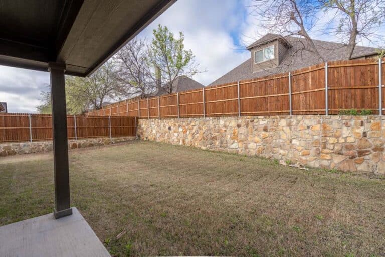 A residential backyard with a stone retaining wall and wooden fence, viewed from a covered patio.