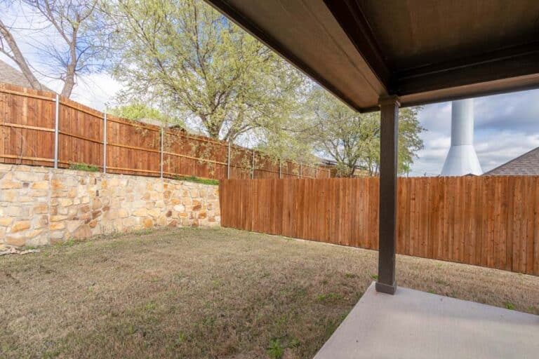 Backyard with a wooden fence and stone retaining wall viewed from under a covered patio.