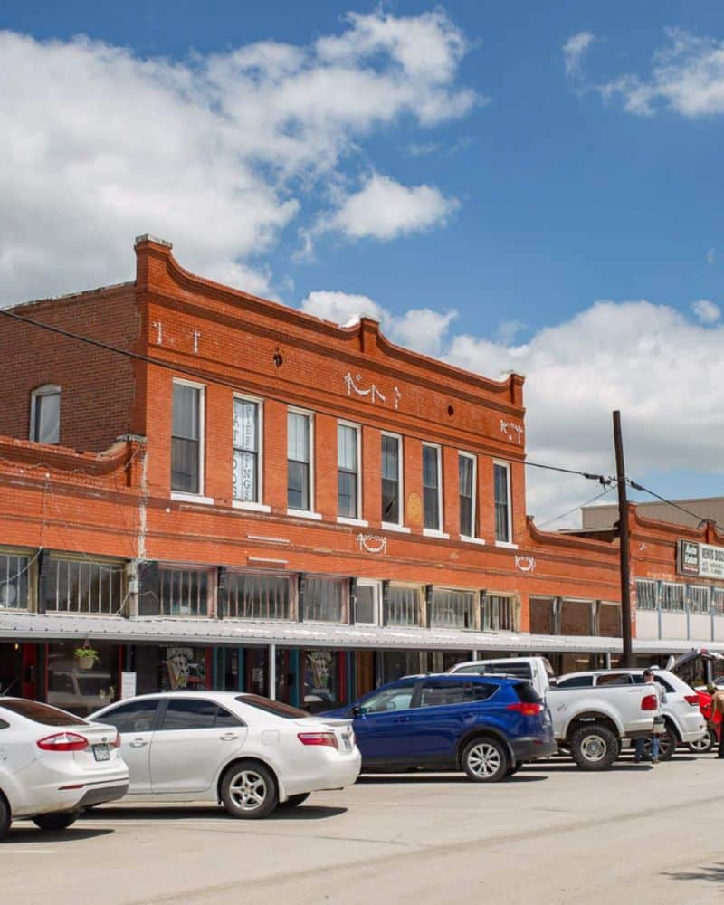 Historic two-story brick building with storefronts and parked cars under a partly cloudy sky.