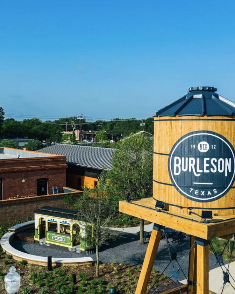 A wooden water tower with "burleson texas" branding stands above a quaint street scene featuring greenery and a yellow bus.
